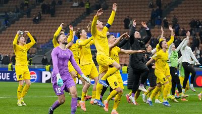 Un equipo de fútbol, con camisetas amarillas, celebra una victoria en el campo, saltando y mostrando euforia. El portero, con uniforme morado, también participa en la celebración, con un estadio lleno de público como telón de fondo.