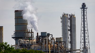 Complejo industrial con chimenea humeante, torres metálicas y red de tuberías bajo un cielo claro. Se observa vegetación en primer plano y una torre de celosía a la derecha. Complejo industrial con chimenea humeante, torres metálicas y red de tuberías bajo un cielo claro. Se observa vegetación en primer plano y una torre de celosía a la derecha.