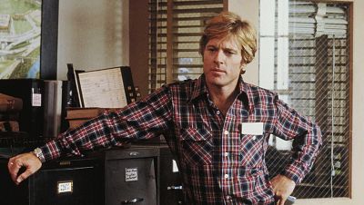 Plano medio de Robert Redford como Henry Brubaker en 1980, con camisa de cuadros y expresión seria junto a un archivador.