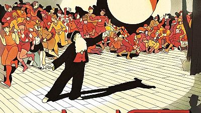 Portada de cómic con una multitud de figuras en tonos rojos y amarillos reunidas en un escenario de madera. Un hombre con barba y traje negro gesticula en primer plano, posiblemente dirigiendo algo.