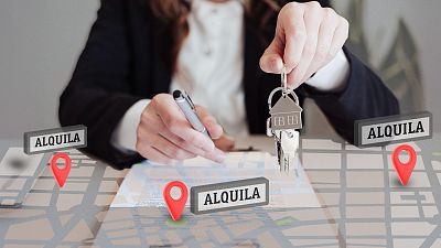 Agente inmobiliario firma documento, sujetando llaves y bolígrafo. Mapa con marcadores rojos 'ALQUILA' sobre la mesa indica gestión de alquileres.