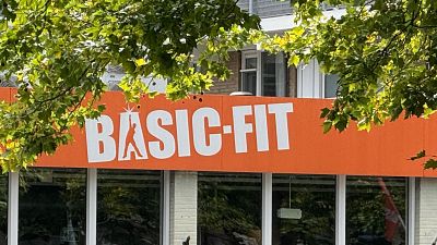 Un letrero naranja de "BASIC-FIT" con letras blancas y una "A" estilizada con una figura en movimiento, se muestra en la fachada de un edificio, parcialmente cubierto por vegetación. Un letrero naranja de "BASIC-FIT" con letras blancas y una "A" estilizada con una figura en movimiento, se muestra en la fachada de un edificio, parcialmente cubierto por vegetación.