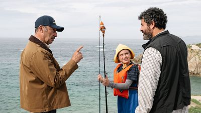 Un fotograma de una película muestra a tres personas en la costa. Un hombre con gorra y chaqueta señala, un niño con sombrero y chaleco sostiene una caña de pescar, y otro hombre con barba observa. Un fotograma de una película muestra a tres personas en la costa. Un hombre con gorra y chaqueta señala, un niño con sombrero y chaleco sostiene una caña de pescar, y otro hombre con barba observa.