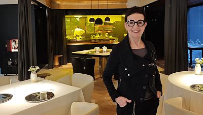 Chef Carme Ruscalleda, en sala de restaurante, viste chaqueta negra y gafas; sonríe a cámara. Ambiente elegante con mesas y cocina visible. Chef Carme Ruscalleda, en sala de restaurante, viste chaqueta negra y gafas; sonríe a cámara. Ambiente elegante con mesas y cocina visible.
