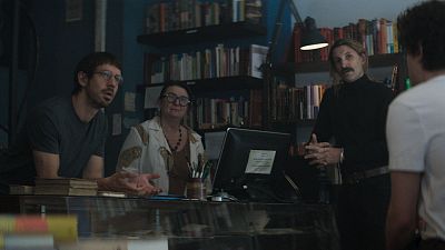 En una librería, un hombre con gafas y barba se apoya en un mostrador, mientras una mujer trabaja en un ordenador. Un hombre con bigote gesticula con las manos.