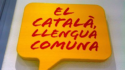 Cartel amarillo en forma de burbuja de diálogo con el mensaje "EL CATALÀ, LLENGUA COMUNA" en letras rojas y gruesas. Cartel amarillo en forma de burbuja de diálogo con el mensaje "EL CATALÀ, LLENGUA COMUNA" en letras rojas y gruesas.