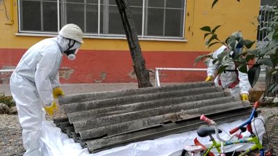 Treballadors amb equips de protecció manipulen plaques de fibrociment ondulades. Al fons, un edifici groc i vermell. A primer pla, una bicicleta infantil i una porteria de futbol. Treballadors amb equips de protecció manipulen plaques de fibrociment ondulades. Al fons, un edifici groc i vermell. A primer pla, una bicicleta infantil i una porteria de futbol.