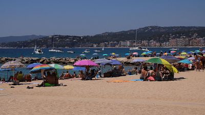 Playa abarrotada en día soleado; gente en la arena, bajo sombrillas, bañándose y embarcaciones en el mar. Ambiente veraniego. Playa abarrotada en día soleado; gente en la arena, bajo sombrillas, bañándose y embarcaciones en el mar. Ambiente veraniego.