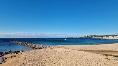 Paisaje costero con playa de arena, mar azul, riachuelo, formaciones rocosas, rompeolas y ciudad en la distancia bajo un cielo despejado. Paisaje costero con playa de arena, mar azul, riachuelo, formaciones rocosas, rompeolas y ciudad en la distancia bajo un cielo despejado.