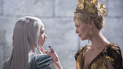 Dos actrices, una con cabello plateado y ropa oscura/plateada, la otra con corona dorada y vestido dorado, interactúan en un interior de piedra.