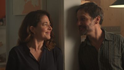 Escena íntima entre un hombre con camisa a rayas y barba, y una mujer con blusa oscura y pendientes, en el interior de una casa.  Ambos sonríen y se miran cerca de un marco de puerta.