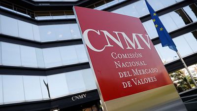 Cartel rojo 'CNMV' en edificio acristalado, con bandera europea cercana.  Probable sede de la Comisión Nacional del Mercado de Valores.
