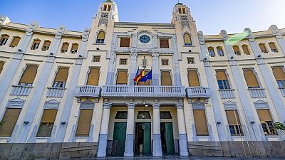 Fachada simétrica de un edificio clásico con dos torres, banderas de España y la UE en un balcón central y rampa de acceso.