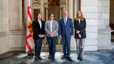 Cuatro personas, dos hombres y dos mujeres en atuendo formal, posan para una foto en un interior con columnas y una bandera.  Contexto político municipal.