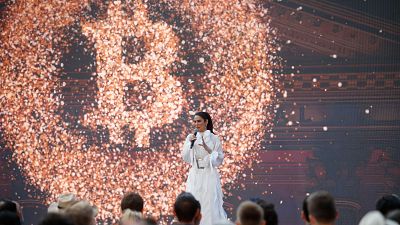 La embajadora, vestida de blanco, habla ante un micrófono con un logo de Bitcoin proyectado detrás, en un evento con audiencia presente.