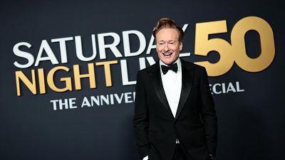 Conan O'Brien sonríe en una foto del 50 aniversario de Saturday Night Live, con el logo del programa visible detrás.