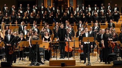 Orquesta y coro en concierto. Director en plataforma central, flanqueado por dos solistas con vestimentas diferentes. Músicos vestidos de negro.