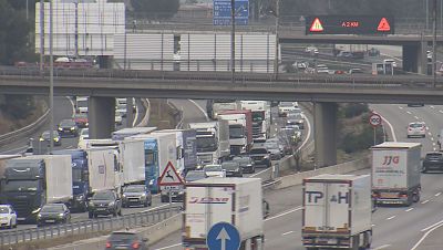 Autopista congestionada con denso tráfico de coches y camiones. Panel informativo a 2 km con señal de advertencia y límite de velocidad de 100 km/h.