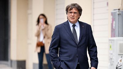 En un plano medio, el político Carles Puigdemont camina de frente por una calle con expresión seria, vistiendo un traje azul marino oscuro y corbata. En un plano medio, el político Carles Puigdemont camina de frente por una calle con expresión seria, vistiendo un traje azul marino oscuro y corbata.