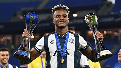 Jugador de fútbol sonriente, con camiseta a rayas, sostiene dos trofeos y una medalla tras una probable ceremonia de premiación.