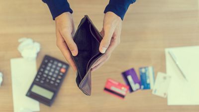 Una cartera de cuero marrón vacía es sostenida por manos, rodeada de objetos como una calculadora, papel arrugado, tarjetas de crédito y un bloc de notas, sugiriendo dificultades económicas. Una cartera de cuero marrón vacía es sostenida por manos, rodeada de objetos como una calculadora, papel arrugado, tarjetas de crédito y un bloc de notas, sugiriendo dificultades económicas.