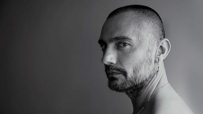 Retrato en blanco y negro de un hombre con cabello oscuro y corto, barba incipiente, pendiente de rayo y cadena, mirando hacia la derecha. Retrato en blanco y negro de un hombre con cabello oscuro y corto, barba incipiente, pendiente de rayo y cadena, mirando hacia la derecha.