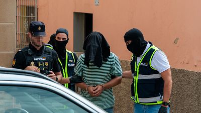 Detención en Melilla: Hombre con rostro cubierto y esposado es rodeado por tres agentes de la Policía Nacional, uno de ellos con cámara. Detención en Melilla: Hombre con rostro cubierto y esposado es rodeado por tres agentes de la Policía Nacional, uno de ellos con cámara.
