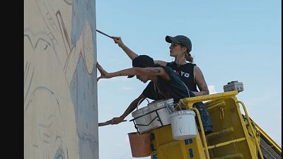 Dos mujeres, Lina Sanabria y Nati Andreoli, pintan un mural desde una grúa amarilla. Utilizan pinceles y se observan cubetas de pintura, con un diseño en tonos claros sobre un fondo azul. Dos mujeres, Lina Sanabria y Nati Andreoli, pintan un mural desde una grúa amarilla. Utilizan pinceles y se observan cubetas de pintura, con un diseño en tonos claros sobre un fondo azul.