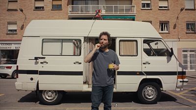 Un hombre con barba y pelo rizado cepilla sus dientes junto a una furgoneta blanca aparcada; la puerta lateral está abierta y se ve una antena en el techo. Un hombre con barba y pelo rizado cepilla sus dientes junto a una furgoneta blanca aparcada; la puerta lateral está abierta y se ve una antena en el techo.