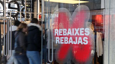 Escaparate de tienda con cartel "REBAIXES REBAJAS" en rojo y blanco. Se aprecian percheros con ropa y personas pasando por delante. Escaparate de tienda con cartel "REBAIXES REBAJAS" en rojo y blanco. Se aprecian percheros con ropa y personas pasando por delante.