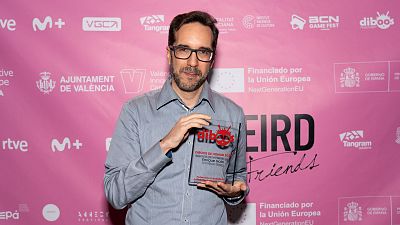 Enrique Gato, con gafas y camisa clara, recibe un premio transparente con un logotipo y una inscripción, posiblemente en un evento. El fondo es de color rosa y presenta varios logotipos. Enrique Gato, con gafas y camisa clara, recibe un premio transparente con un logotipo y una inscripción, posiblemente en un evento. El fondo es de color rosa y presenta varios logotipos.