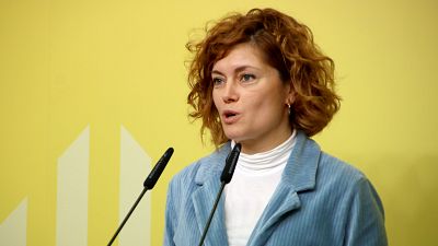 Portavoz de ERC ofreciendo una rueda de prensa. Lleva chaqueta azul y jersey blanco, hablando a dos micrófonos sobre un fondo amarillo con líneas verticales. Portavoz de ERC ofreciendo una rueda de prensa. Lleva chaqueta azul y jersey blanco, hablando a dos micrófonos sobre un fondo amarillo con líneas verticales.