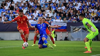 Partido de fútbol: jugador eslovaco (camiseta azul, #7) intenta interceptar el balón mientras un jugador español (camiseta roja, #2) lo conduce. Un portero (verde fluorescente) observa.
