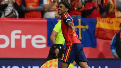 El futbolista español Lamine Yamal, con la camiseta número 19, corre en el césped persiguiendo el balón durante un partido de fútbol ante un rival de blanco. El futbolista español Lamine Yamal, con la camiseta número 19, corre en el césped persiguiendo el balón durante un partido de fútbol ante un rival de blanco.