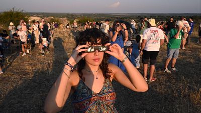 Mujer con gafas de eclipse y pulsera '2026' observa evento astronómico junto a multitud en ladera. Mujer con gafas de eclipse y pulsera '2026' observa evento astronómico junto a multitud en ladera.