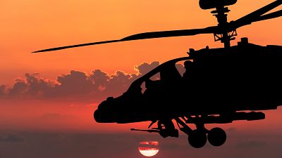 Un helicóptero militar oscuro se recorta contra un cielo crepuscular, con el sol poniente creando un resplandor rojizo y anaranjado. Se distinguen las hélices y el armamento del helicóptero. Un helicóptero militar oscuro se recorta contra un cielo crepuscular, con el sol poniente creando un resplandor rojizo y anaranjado. Se distinguen las hélices y el armamento del helicóptero.