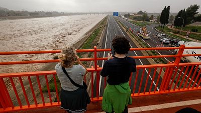 Río desbordado inunda carretera, vehículos detenidos. Dos personas en puente observan la crecida; una graba con móvil.  Magnitud de la inundación.