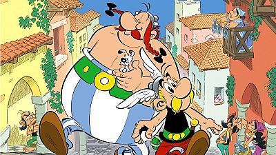 En la portada de un cómic, se destacan dos personajes principales: un hombre pequeño con casco y bigote, y otro corpulento con trenzas, que sostiene un perro. Detrás, se observa un pueblo con edificios de tejados rojos. En la portada de un cómic, se destacan dos personajes principales: un hombre pequeño con casco y bigote, y otro corpulento con trenzas, que sostiene un perro. Detrás, se observa un pueblo con edificios de tejados rojos.