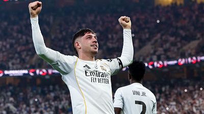 Fede Valverde celebra un gol con los brazos levantados, vistiendo la camiseta blanca de su equipo, con el logo de un patrocinador. Se observa a otro jugador de espaldas y un estadio lleno de público.