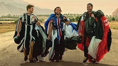 Tres paracaidistas, sonrientes y con cascos, posan en un camino montañoso. Uno de ellos viste un traje con los colores de una bandera, mientras que los otros dos llevan trajes de colores contrastantes.