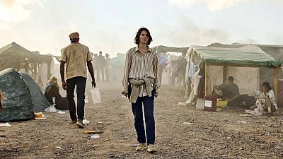 Mujer caminando hacia la cámara en un campamento pobre; un hombre se aleja. Ropa sencilla, ambiente desolado. Contrastes sociales. Mujer caminando hacia la cámara en un campamento pobre; un hombre se aleja. Ropa sencilla, ambiente desolado. Contrastes sociales.