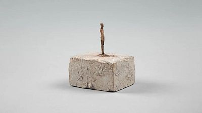 Escultura estilizada marrón oscuro, figura delgada y alargada sobre pedestal rectangular gris claro de textura rugosa. Fondo gris uniforme. Escultura estilizada marrón oscuro, figura delgada y alargada sobre pedestal rectangular gris claro de textura rugosa. Fondo gris uniforme.