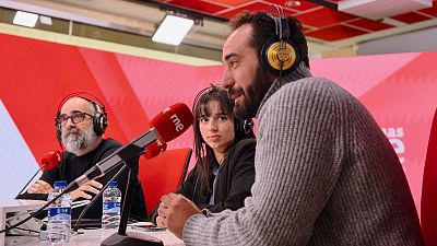 Tres personas, dos hombres y una mujer, se encuentran en un estudio de radio frente a micrófonos. Los hombres llevan auriculares y barba, mientras que la mujer tiene el pelo oscuro y flequillo. Hay botellas de agua sobre la mesa.