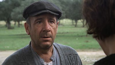 Alfredo Landa, con gorra oscura y jersey gris, posa con expresión preocupada. El fondo revela un paisaje rural con vegetación, posiblemente durante la filmación de una película. Alfredo Landa, con gorra oscura y jersey gris, posa con expresión preocupada. El fondo revela un paisaje rural con vegetación, posiblemente durante la filmación de una película.