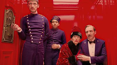 Cuatro personajes de 'El Gran Hotel Budapest': Adrien Brody, Tony Revolori ("LOBBY BOY"), Tilda Swinton y Ralph Fiennes, posan en un interior rojo vibrante.