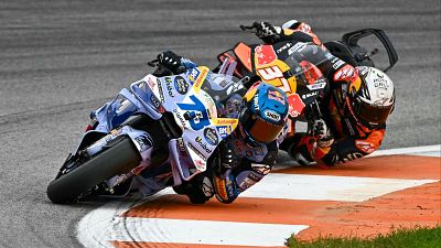 Dos pilotos de MotoGP compiten en una curva del circuito. Uno lleva una moto azul y blanca con el número 73, mientras que el otro pilota una moto naranja, negra y blanca.