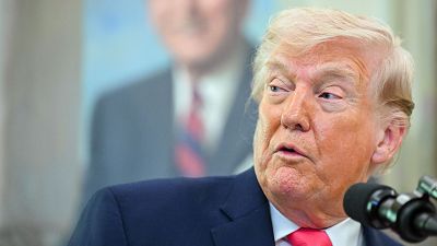 Donald Trump, con traje azul y corbata roja, está de pie frente a un micrófono. Se encuentra en un espacio donde se distingue la imagen borrosa de otro hombre vestido formalmente. Donald Trump, con traje azul y corbata roja, está de pie frente a un micrófono. Se encuentra en un espacio donde se distingue la imagen borrosa de otro hombre vestido formalmente.