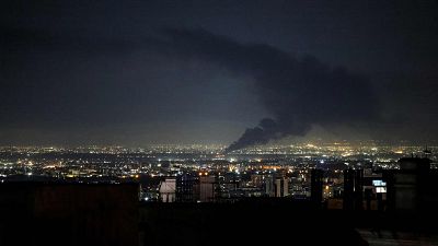 Una ciudad iluminada por la noche, con una columna de humo negro ascendiendo hacia el cielo. Las luces de la ciudad contrastan con la oscuridad y el humo, sugiriendo un evento reciente. Una ciudad iluminada por la noche, con una columna de humo negro ascendiendo hacia el cielo. Las luces de la ciudad contrastan con la oscuridad y el humo, sugiriendo un evento reciente.