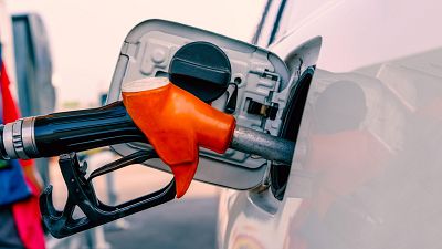 Una manguera de gasolina con boquilla naranja y mango negro se encuentra conectada al depósito de un coche blanco, posiblemente en un contexto económico relacionado con el precio de los combustibles.