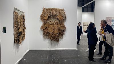 Grupo de personas observando obras de arte, posiblemente tapices de fibras naturales, en una sala de exposiciones. Un hombre explica las piezas a otros asistentes. Grupo de personas observando obras de arte, posiblemente tapices de fibras naturales, en una sala de exposiciones. Un hombre explica las piezas a otros asistentes.